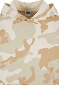 Beige camo hettegenser laget av mykt stoff. Har en justerbar hette og uregelmessige mønstre i lyse tan- og kremnyanser.