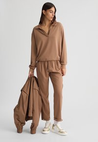 Completo loungewear marrone con un pullover e pantaloni a vita elastica. Texture liscia, vestibilità comoda. Una giacca marrone coordinata è tenuta in una mano.
