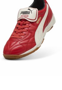 Crvena kožna nogometna cipela s bijelim detaljima, s podstavljenim ovratnikom, vezanjem i gumenom potplatom. Logotip marke na jeziku.