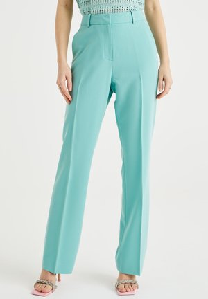 Vrouw draagt een turquoise broek met hoge taille en rechte pijpen, gecombineerd met een licht gebreid cropped top en roze open-toe hakken met gevlochten bandjes.