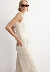 Mujer con gafas de sol oscuras, top de punto color crema sin mangas y pantalones anchos de cintura alta en beige, con la mano en el bolsillo trasero.