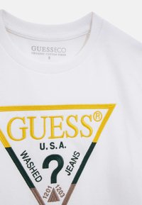 Weißes T-Shirt aus Bio-Baumwolle mit einem mehrfarbigen gestickten dreieckigen Logo, das "GUESS", "U.S.A." und "WASHED JEANS" zeigt.