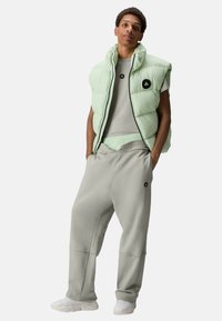 Lichtgroene gewatteerde bodywarmer met een hoge kraag en een zwart logo, gecombineerd met grijze joggingbroek. De outfit heeft een comfortabele pasvorm en een zachte textuur.