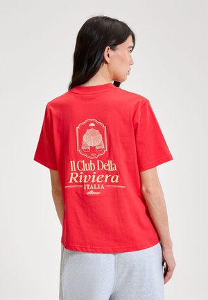 Ženska z dolgimi temnimi lasmi, oblečena v rdečo majico z bež napisom in grafiko, "Il Club Della Riviera Italia," stoji obrnjena stran.