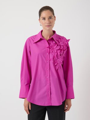 Rosa skjorta med knappar och en asymmetrisk blommotiv-applikation på ena axeln. Långa ärmar, kragdesign och mjuk textur.