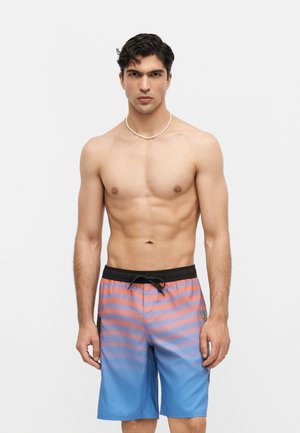 Mladý muž s krátkymi tmavými vlasmi, oblečený v modro-ružové pruhované plavky a biely náhrdelník, stojaci na bielom pozadí.