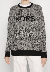 Suéter de manga larga con estampado de leopardo en negro y gris, con el logo "KORS" en negro, usado sobre una camisa blanca y combinado con pantalones blancos.