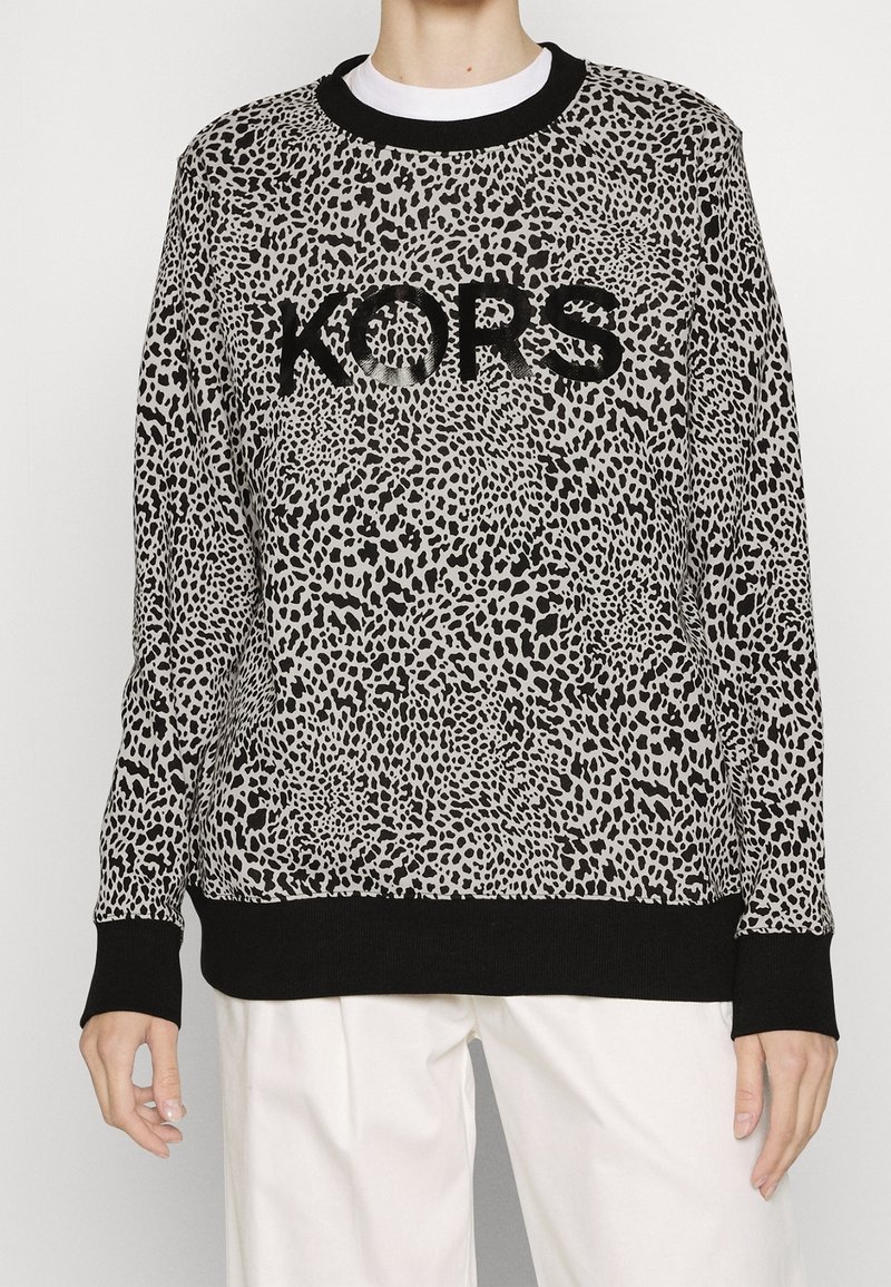 Suéter de manga larga con estampado de leopardo en negro y gris, con el logo "KORS" en negro, usado sobre una camisa blanca y combinado con pantalones blancos.