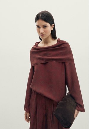 Femme aux cheveux raides et foncés porte une robe ample bordeaux et tient une pochette souple marron foncé sur un fond clair uni.