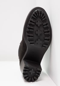 Schwarze Stiefelsohle mit tiefen Traktionsprofilen und kreuzförmigen Mustern, fotografiert vor einem weißen Hintergrund.