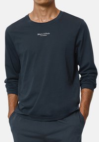 Man wearing a dark navy long-sleeve shirt with "Marc O'Polo" text on the chest, hands in matching dark navy pants pockets.