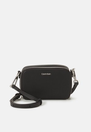 Schwarze rechteckige Umhängetasche mit silbernem Reißverschluss und Verschluss, Calvin Klein Logo vorne, verstellbarer Riemen vorne aufgerollt vor weißem Hintergrund.