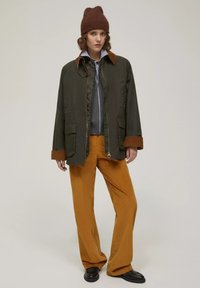 Olivegrüne Jacke mit einem braunen Cordkragen, kombiniert mit orangen Cordhosen. Das Model trägt eine braune Strickmütze und einen grauen Pullover darunter.