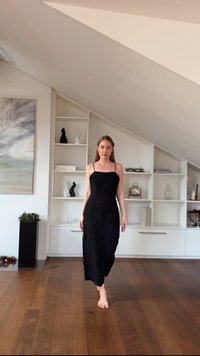 Schwarzes plissiertes Maxikleid mit dünnen Trägern, tailliertem Oberteil und fließendem Rock. Holzboden und minimalistische Regale im Hintergrund.