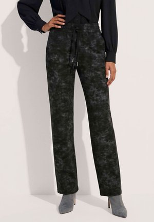 Vrouw draagt zwarte, met patroon en trekkoord voorzien broek, zwarte blouse met lange mouwen en grijze enkellaarzen met hoge hak.