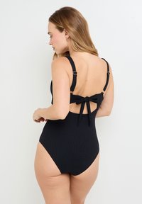 Maillot de bain une pièce noir en tissu texturé, avec des bretelles réglables et un détail en nœud au dos, dotée d'un décolleté dans le dos.