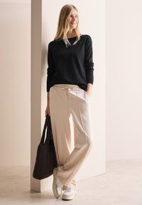 Schwarzer Strickpullover, beiger weiter Jogginghose mit Kordelzug, weiße Sneakers und eine schwarze Tragetasche, vor einem neutralen Hintergrund.