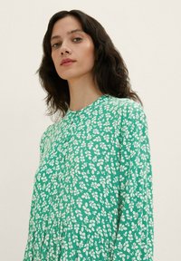 Grünes Kleid aus leichtem Stoff, mit einem weißen Blumenmuster, langen Ärmeln, einem runden Ausschnitt und Knopfdetails an der Vorderseite.