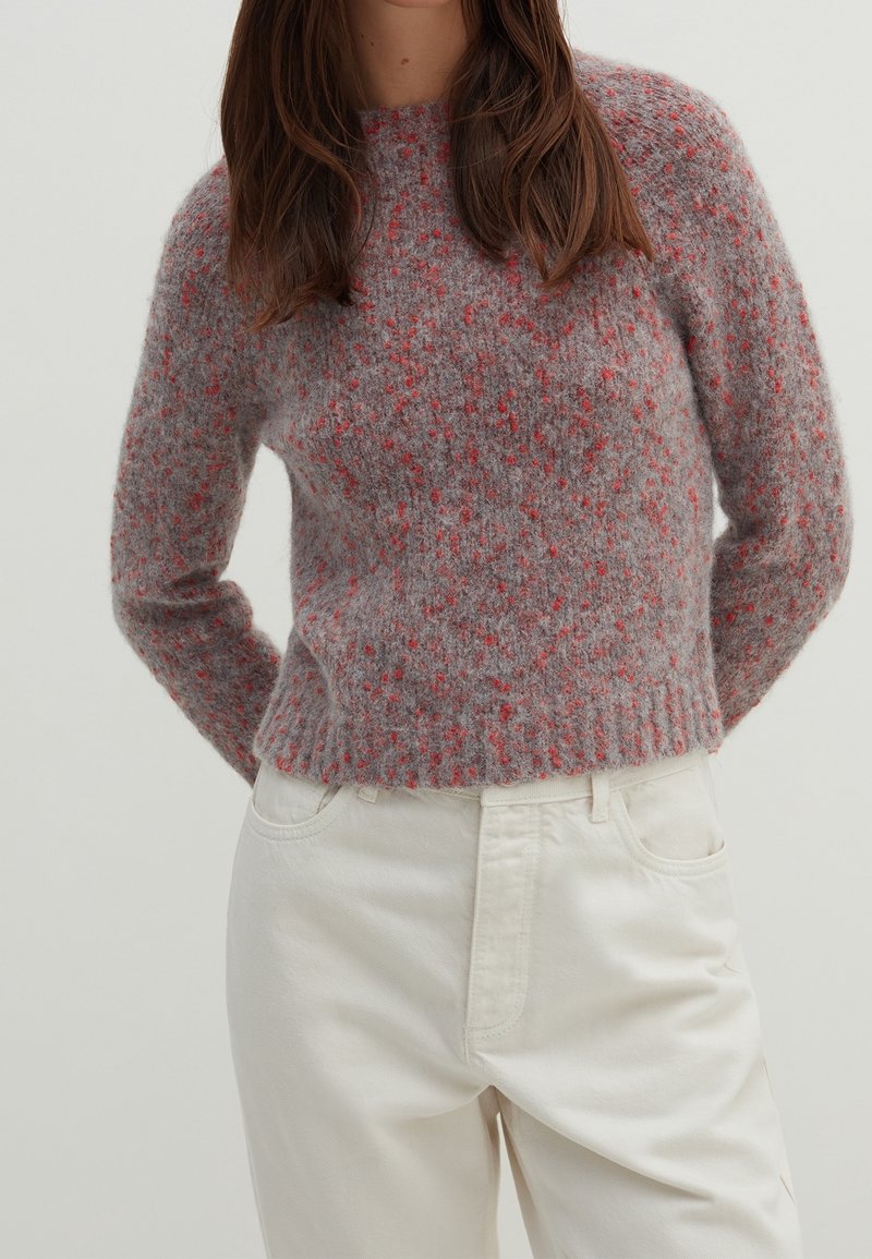 Woman wearing a fuzzy gray sweater with red speckles and off-white high-waisted pants, standing with hands behind her back.