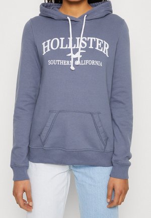 Person trägt einen blauen Hollister-Hoodie mit weißen Kordeln und dem Schriftzug "Southern California", kombiniert mit zweifarbigen Jeans in Hellblau und Weiß.