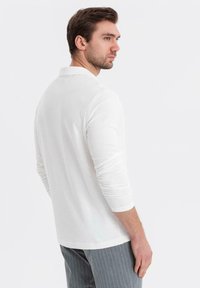 Man med kort brunt hår, iförd en vit långärmad polo och grå ränderade byxor, tittar över sin axel åt höger.