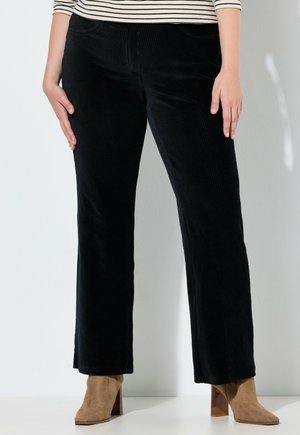 Donna che indossa pantaloni in corduroy neri e stivaletti in suede marroni, in piedi con le mani appoggiate sulle cosce su uno sfondo bianco.
