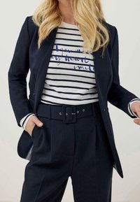 Blazer bleu marine sur une chemise blanche à rayures bleu marine avec un texte graphique bleu. Pantalon taille haute avec une large ceinture et des poches visibles.