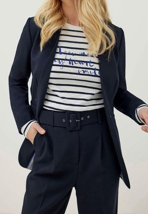 Blazer bleu marine sur une chemise blanche à rayures bleu marine avec un texte graphique bleu. Pantalon taille haute avec une large ceinture et des poches visibles.