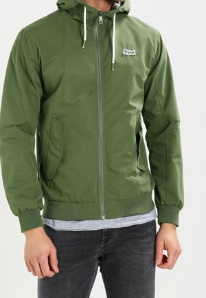 Homme portant une veste à capuche verte zippée avec des cordons blancs et des poches latérales sur un t-shirt gris et un jean noir.