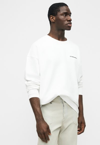 Άνδρας με άσπρο πουκάμισο μακρυμάνικο με μικρό μαύρο κείμενο στο στήθος, μπεζ παντελόνι, κοιτάζει δεξιά του μπροστά σε ένα απλό ανοιχτό φόντο.