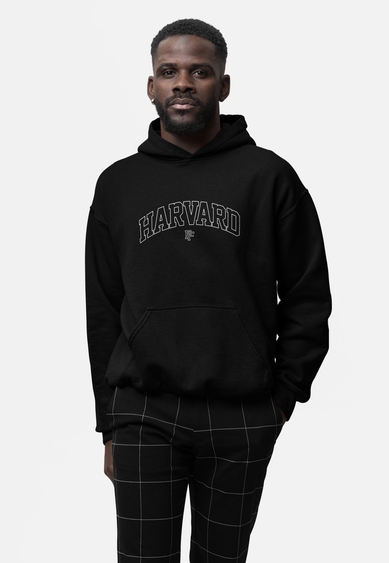 Man standing facing forward, wearing a black Harvard hoodie and black pants with white grid lines, neutral expression, plain light background.
