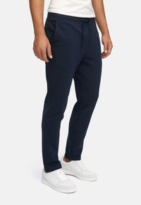 Mand iført navyblå snorebukser og hvide sneakers, stående mod en helt hvid baggrund.