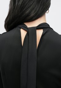 Fekete szövetszövet felsőrész hátsó részén masnival megkötve, kulcslyuk kivágással. Sima textúra és puha, légies kialakítás.
