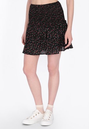 Zwarte minirok met rood en roze bloemenprint, smock taille, gedragen met witte sneakers en doorzichtige enkelsokken op blote benen.