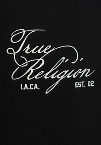 Fekete szövet, amelyen fehér hímzett szöveg található: "True Religion", díszes, íves betűtípussal. Alatta szerepel az "L.A.CA." és az "EST. 02".