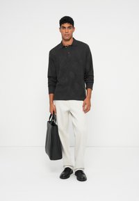 Schwarzes Langarm-Polo mit dezentem Texturmuster, kombiniert mit heller Hose und einer schwarzen Tragetasche mit eleganten Griffen und minimalistischem Design.