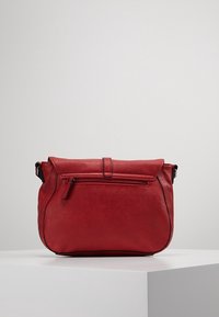 Červená kožená taška s klopou a předním kapsou na zip. Má zaoblené hrany a nastavitelný popruh pro nošení přes rameno.