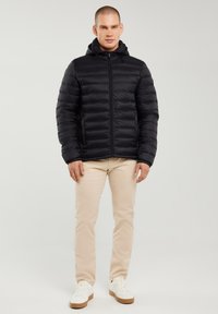 Hombre de pie con una chaqueta acolchada negra con capucha, pantalones beige y zapatillas blancas, sobre un fondo claro y liso.