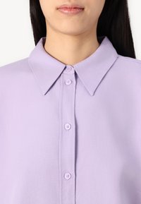Lilla skjorte med knapper som har en klassisk krage, teksturert stoff, og fem matchende knapper langs frontplagget.