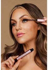 En mascaratube med matt rosa finish hålls av en person som applicerar den, med en svart borste och klara borst för applicering.