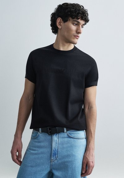 Czarny bawełniany t-shirt z krótkim rękawem, z teksturowanym czarnym logo na przodzie. Noszony z jasnoniebieskimi jeansami i czarnym paskiem.