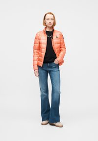 Femme portant une veste matelassée orange sur un chemisier noir, jean bleu évasé, chaussures beiges, debout avec une main dans la poche.