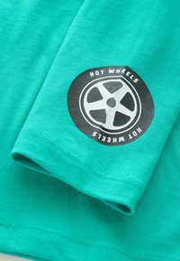 Tirkīza krāsas kokvilnas krekls ar melnu apaļu logo, kurā redzams riteņu dizains un uz piedurknes baltiem burtiem rakstīts "HOT WHEELS".