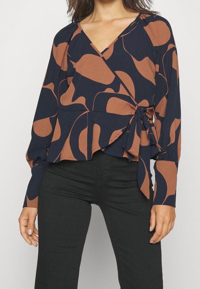 Femme portant un chemisier cache-cœur floral noir et marron à manches longues et un pantalon noir, noué à la taille avec un nœud sur le côté.