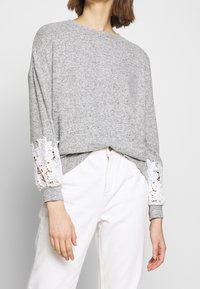 Grå sweatshirt med långa ärmar som har vit spetsdetalj vid ärmsluten, ihopparad med vita byxor i ett mjukt, texturerat tyg.