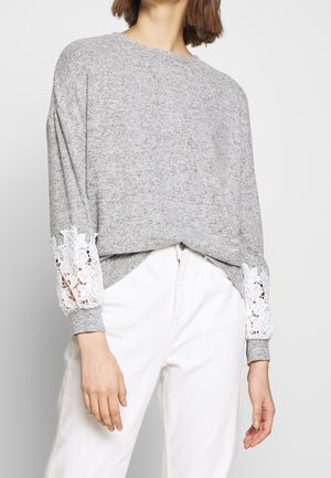 Grauer Sweatshirt mit langen Ärmeln und weißen Spitzenbesätzen an den Manschetten, kombiniert mit weißen Hosen aus einem weichen, strukturierten Stoff.