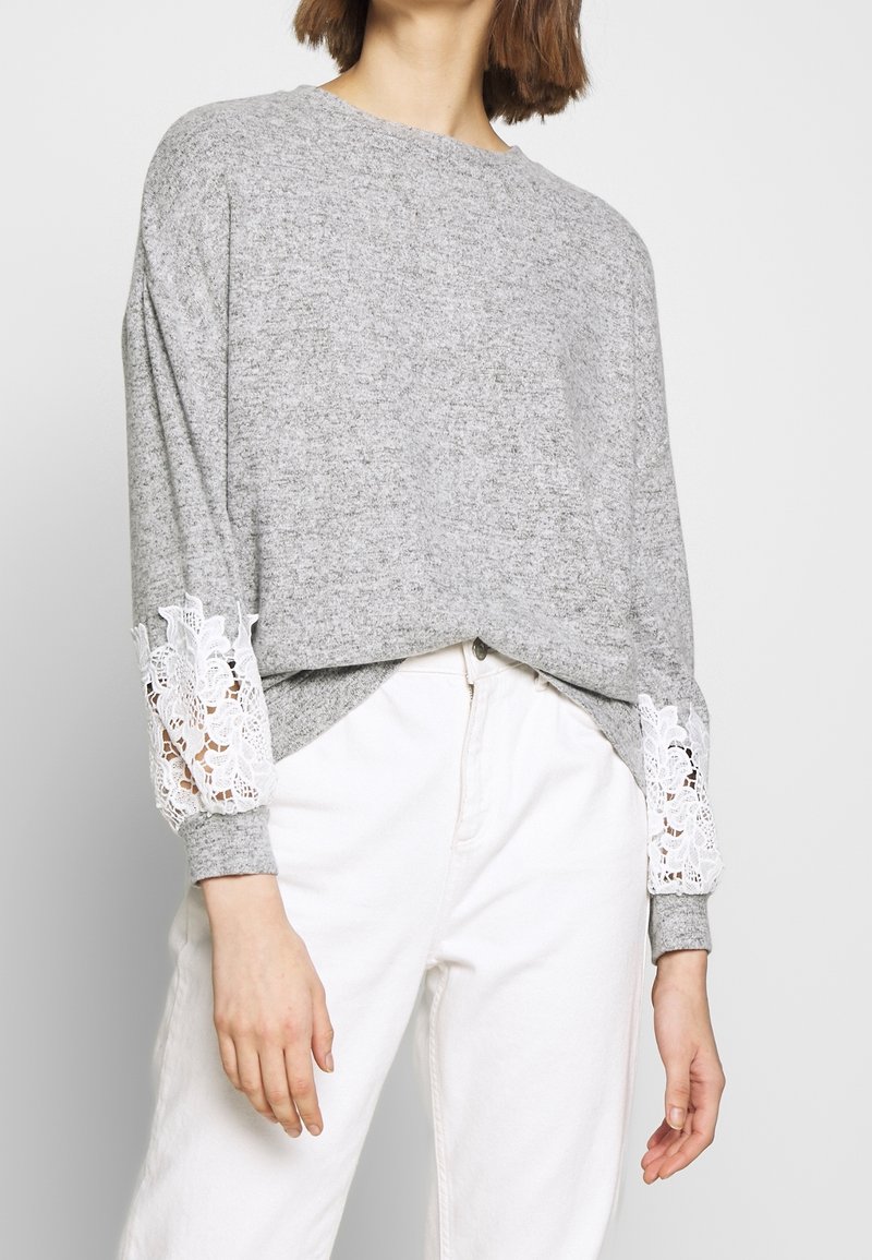 Grå sweatshirt med långa ärmar som har vit spetsdetalj vid ärmsluten, ihopparad med vita byxor i ett mjukt, texturerat tyg.