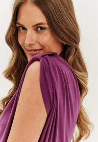 Fialová bezrukávová blúzka s nariasenými detailmi na pleciach a jemnou, splývavou textúrou. Nosená s voľnými, vlnitými vlasmi rámujúcimi tvár.