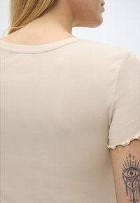 Γυναίκα με ξανθά μαλλιά που φοράει μπεζ κοντομάνικο πουλόβερ με ριμπ ύφανση και τελείωμα σαλάτας, εμφανίζοντας μαύρο μάτι και τατουάζ στο χέρι της.