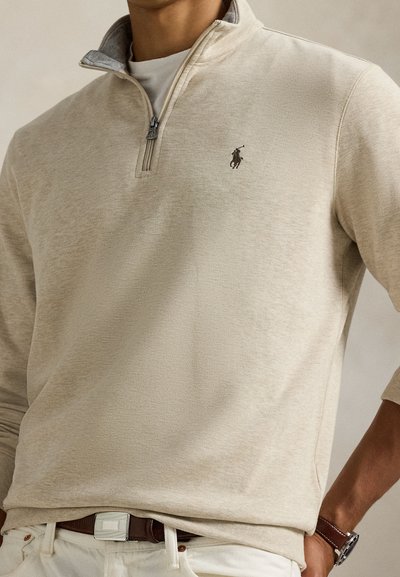 Beige quarter-zip genser laget av mykt stoff, med en grå krage og en liten mørk logodetalj. Lange ermer med avslappet passform.